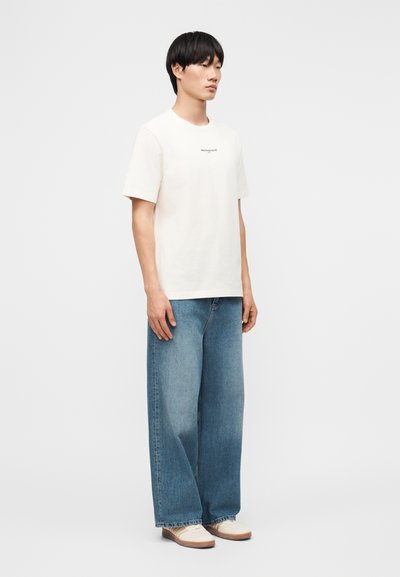 Άνδρας στέκεται φορώντας λευκό κοντομάνικο πουκάμισο με κείμενο, φαρδιά μπλε τζιν παντελόνια και λευκά παπούτσια μπροστά σε απλό γκρι φόντο.