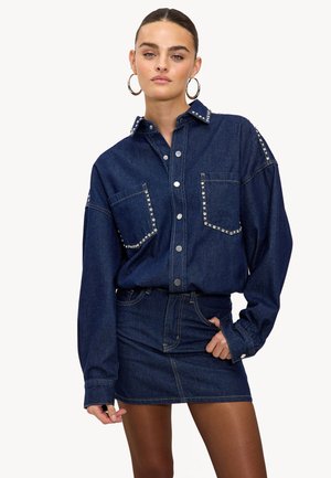 Donkerblauwe denimshirt met twee borstzakken, zilveren studs, knoopsluiting aan de voorkant en lange mouwen, gecombineerd met een bijpassende denim minirok.