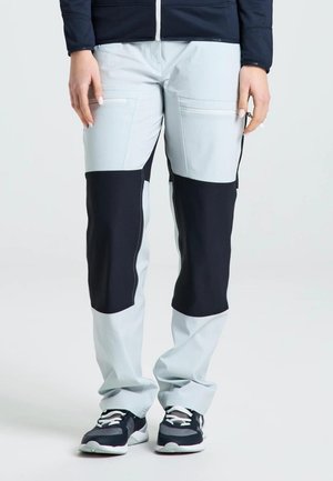 Una persona luce unos pantalones gris claro con paneles oscuros en las rodillas y bolsillos con cremallera, una chaqueta oscura y zapatillas negras sobre un fondo liso.