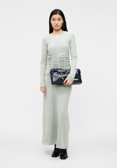 Dolga zelena obleka s teksturo nabranega materiala, kombinirana s črnim usnjenim spaklom z zaponko in obeskom v obliki ključa. Črni gležnjarji.