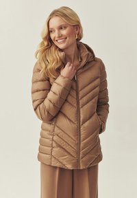 Veste matelassée beige avec un motif en chevrons, fermeture éclair et capuche. Le matériau est lisse et légèrement brillant, avec des poches latérales.