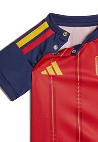 Sarkans un tumši zils sporta krekls ar dzeltenām un sarkanām svītrām uz piedurknēm, dzeltenais Adidas logo, presējamie pogas apkaklē un vertikāli sarkani un dzelteni akcenti.