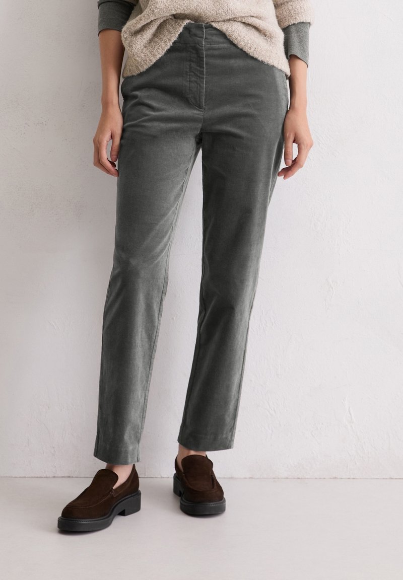 Personne portant un pantalon gris droit, un pull beige et des mocassins marron foncé, debout contre un mur blanc.