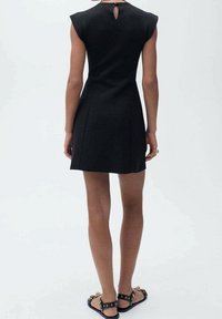 Robe noire à manches courtes, design ajusté, longueur au genou, détail en forme de trou clé au dos et texture lisse. Portée avec des sandales plates.