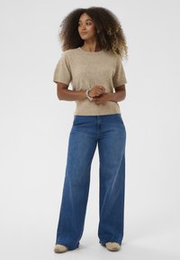 Maglione beige a maniche corte con scollatura rotonda, abbinato a jeans blu a gamba larga e scarpe beige, su uno sfondo bianco semplice.