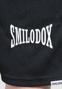 Svarta rippade shorts med en framträdande vit "SMILODOX"-logotyp placerad diagonalt. Tyget har en strukturerad yta.
