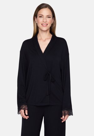 Frau mit langen braunen Haaren, die ein schwarzes Wickeloberteil mit Spitzenmanschetten und passende schwarze Hose trägt, lächelt vor einem schlichten weißen Hintergrund.