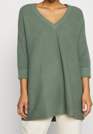 Person trägt einen locker sitzenden, strukturierten grünen V-Ausschnitt-Pullover mit dreiviertel Ärmeln und hellen Hosen.