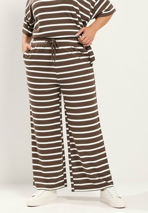 Persona que lleva pantalones anchos marrones con rayas horizontales blancas y camiseta a juego, combinados con zapatillas blancas, de pie con las manos en los bolsillos.