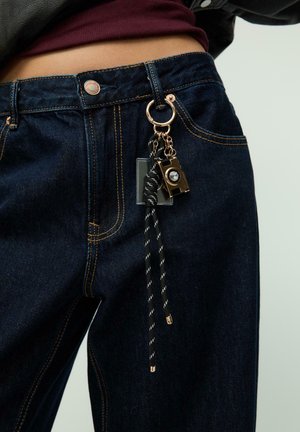 Tmavě modré džíny z denimu se zlatým prošíváním a dekorativním přívěskem na klíče. Přívěsek zahrnuje malou černou kameru a pruhovanou šňůru.