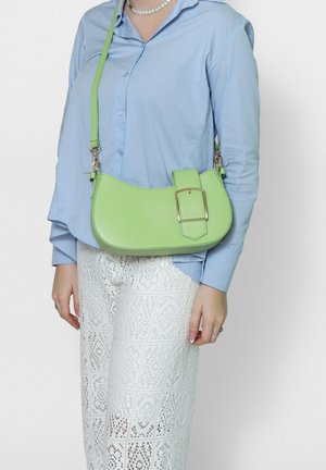 Donna che indossa una camicia azzurra con bottoni e pantaloni bianchi in pizzo, con una piccola borsa a tracolla verde chiaro con una grande fibbia.