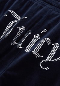 Navi modra velur tkanina z okrasnimi kamenčki, ki tvorijo napis "Juicy." Gladka tekstura z izrazitim sijajem draguljev.