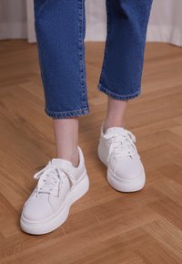 Baskets synthétiques blanches avec un bout rond, une semelle texturée et un système de laçage, assorties à un pantalon en denim bleu cropped, sur un sol en bois.