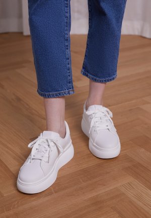 Zapatillas sintéticas blancas con punta redonda, suela texturizada y diseño de cordones, combinadas con pantalones de mezclilla azul recortados, sobre un suelo de madera.