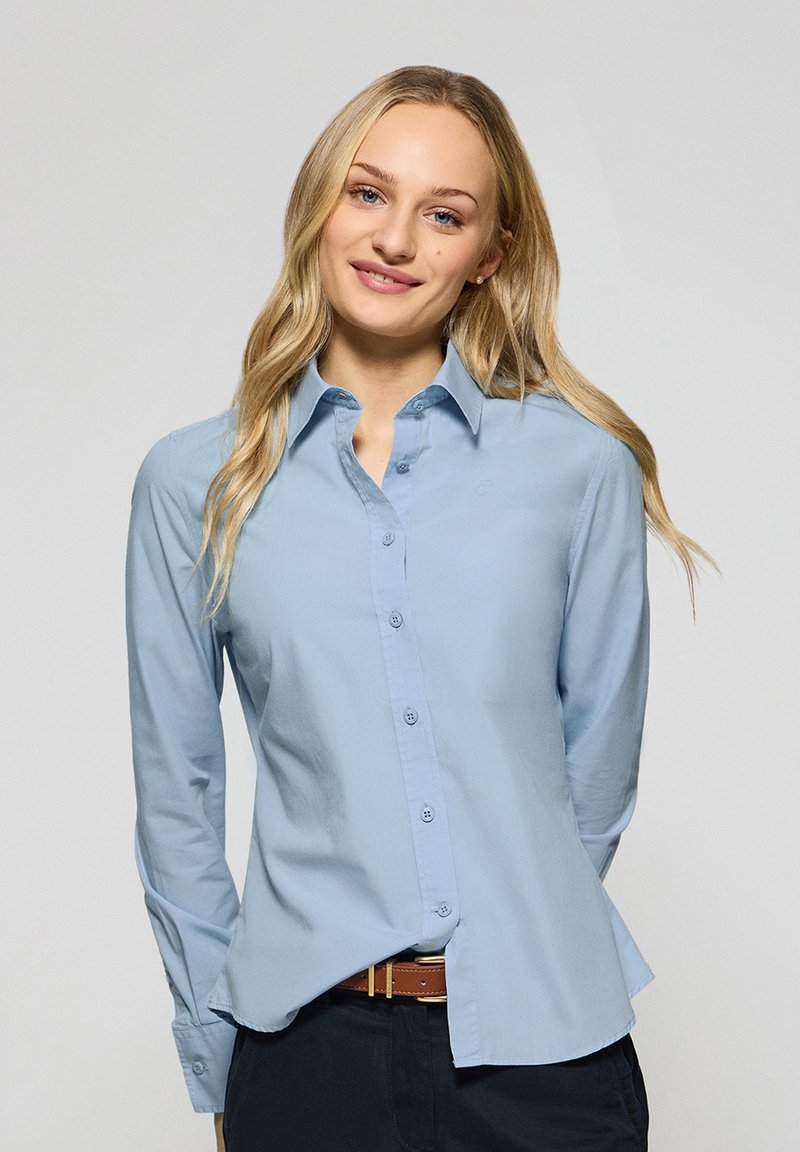 Ljusblå skjorta med knappar, lång ärm, skräddarsydd passform, mjuk textur och rundad nederkant. Har krage och subtil logotyp på bröstet.