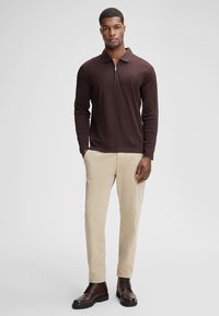 Braunes langärmliges Polo-Shirt mit einem Reißverschlusskragen, kombiniert mit beigen Hosen und braunen Stiefeln, bietet eine lockere Passform und eine glatte Stofftextur.