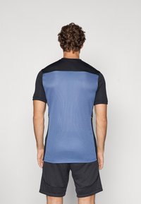 Kortsleppet idrettsgenser med blå mesh-bakstykke, svarte skulderpaneler og gule detaljer på sidene. Båret med svarte shorts.