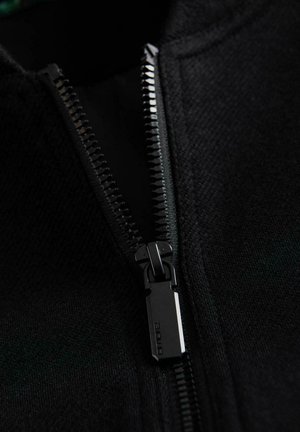 Vêtements noirs à fermeture éclair mettant en valeur un tissu texturé, disposant d'une fermeture éclair en métal durable avec un onglet rectangulaire et des détails de Couture serrés.