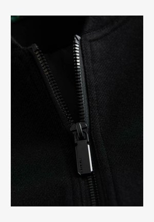 Vêtements noirs à fermeture éclair mettant en valeur un tissu texturé, disposant d'une fermeture éclair en métal durable avec un onglet rectangulaire et des détails de Couture serrés.