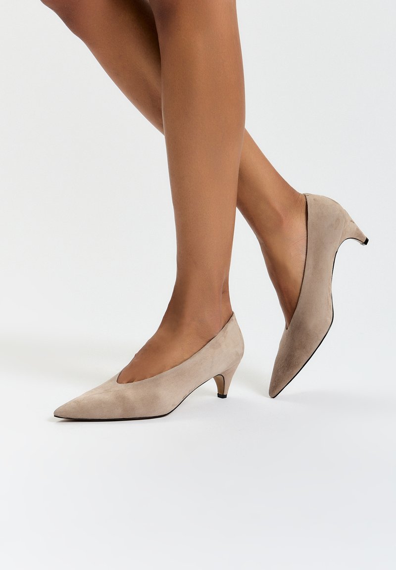 Décolleté in camoscio beige con punta affilata, tacco basso a kitten e ritagli laterali. Superficie texture e dettagli sulla suola nera.
