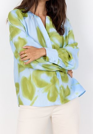 Femme portant une chemise à manches longues tie-dye bleu clair et vert, avec un pantalon de couleur crème, bras croisés montrant des bagues en or.