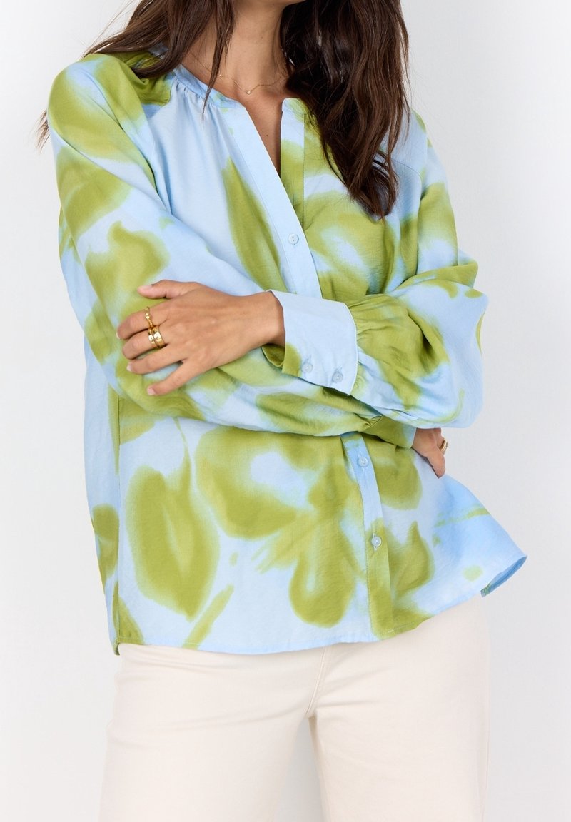 Femme portant une chemise à manches longues tie-dye bleu clair et vert, avec un pantalon de couleur crème, bras croisés montrant des bagues en or.