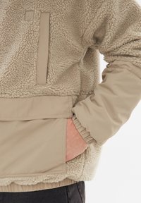 Beige fleecejakke med struktureret finish, der har en frontlomme og elastiske manchetter. Glat stof ved kanten og detaljering på brystet.