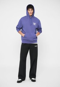 Πορτοκαλί φούτερ με μπροστινή τσέπη και λευκό λογότυπο, σε συνδυασμό με μαύρα παντελόνια φαρδιάς γραμμής. Λείο ύφασμα με άνετη εφαρμογή.