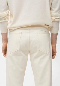 Pullover a maglia crema sopra pantaloni beige chiari con due tasche posteriori. Il tessuto appare morbido, con una finitura leggermente testurizzata.