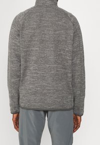 Veste en polaire grise avec un col montant, surface texturée et manches raglan. Présente un ourlet lisse et est portée avec un pantalon gris.