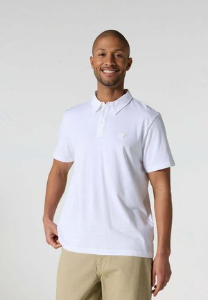 Hombre sonriente, con camiseta polo blanca de manga corta y pantalones beige, posando frente a un fondo liso de color gris claro.