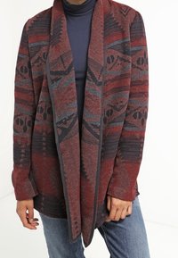 Long cardigan ouvert avec des motifs géométriques en rouge, gris et noir porté sur un col roulé sombre et un jean bleu.