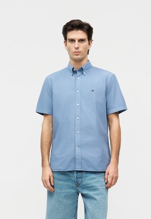 Νέος άνδρας με σκούρα μαλλιά, που φοράει ανοιχτό μπλε κοντομάνικο πουκάμισο με κουμπιά και ανοιχτό μπλε τζιν, στέκεται μπροστά σε ένα απλό φόντο.