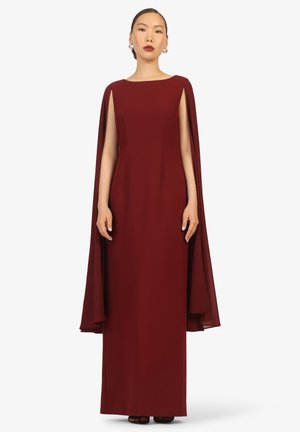 Woman standing wearing a floor-length, sleeveless burgundy gown with attached flowing cape and gold hoop earrings, against white background.