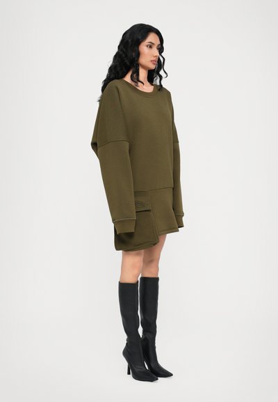 Olivovo zelené oversized mikinové šaty s dlhými rukávmi, so bočnou kapsou a nenápadným brandingom. Skombinované s čiernymi čižmami s vysokým podpätkom po kolená.