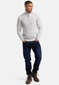 Hellgrauer Kabelstrickpullover mit Stehkragen, kombiniert mit dunkelblauen Jeans und braunen Schnürstiefeln. Texturiert und tailliert.