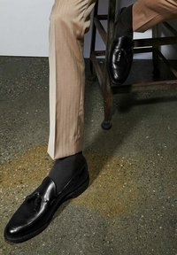 Schwarze Lederloafers mit Quasten, getragen mit dunklen Socken und beige gestreiften Hosen, auf einem strukturierten Betonboden platziert.