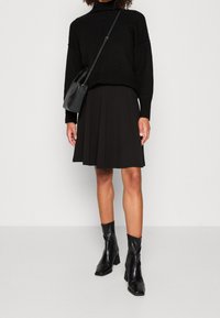 Černý, převeliký svetr s vysokým límcem, zkombinovaný s černou skladanou sukní. Černé kotníkové boty se čtveratou špičkou. Malá černá kabelka.