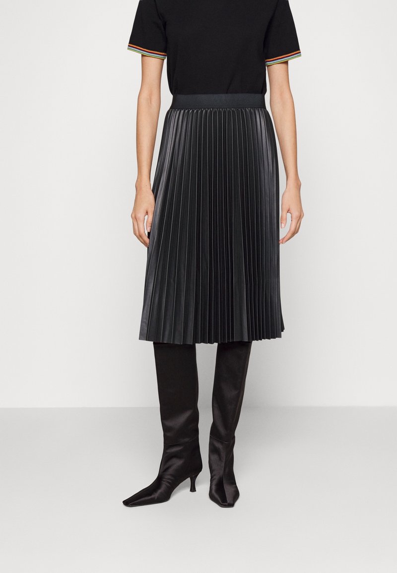 Femme vêtue d'une jupe midi plissée noire, d'un haut noir avec des bords de manches rayés et des bottines noires à bout pointu et talons, debout sur fond blanc.