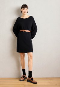 Robe en tricot noir avec des manches surdimensionnées, cintrée par une ceinture en cuir marron. Chaussures marron avec découpes, associées à des chaussettes côtelées noires.