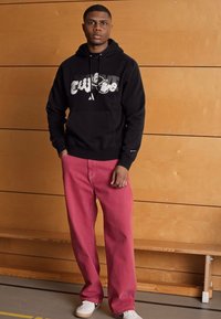 Felpa nera con design grafico bianco, abbinata a pantaloni larghi rosa. Sneakers bianche completano l'outfit casual. Parete di legno sullo sfondo.