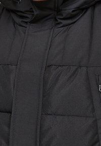 Svart vadderad jacka med en slät textur, strukturerad design och en framszipper med en avtagbar huva. Anmärkningsvärda sömnadsdetaljer.