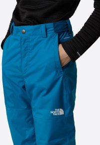 Modré voděodolné lyžařské kalhoty mají pevný pas, boční kapsy a logo značky The North Face na spodní části nohavice.