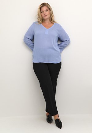 Blouse bleu clair à manches longues et en V avec une poche sur le devant, associée à un pantalon noir ajusté et des escarpins noirs à bout pointu.