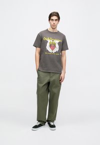 Joven de pie vistiendo una camiseta gráfica oscura con texto amarillo y pantalones verde oliva, complementados con zapatillas negras y blancas.