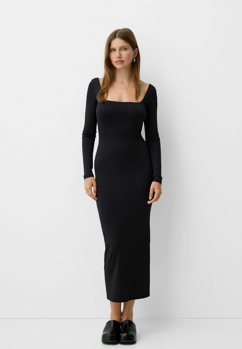 Donna che indossa un lungo abito aderente nero con scollatura quadrata e maniche lunghe, in piedi davanti a uno sfondo bianco uniforme.