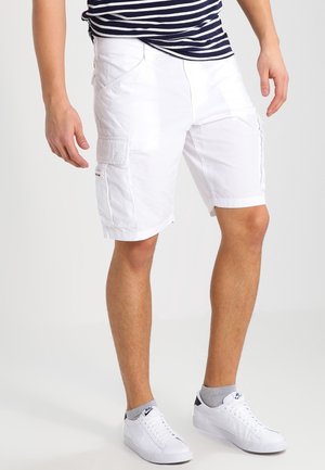 Mężczyzna w białych spodenkach cargo, pasiastym granatowo-białym t-shircie, szarych skarpetkach i białych trampkach Nike stoi na białym tle.