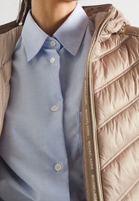 Chaqueta acolchada color beige claro con cierre de cremallera y detalle del logotipo en la cremallera, superpuesta a una camisa azul con cuello y botones blancos.