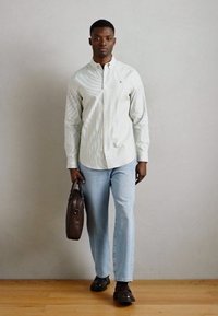 Λευκό πουκάμισο με κάθετες ανοιχτό μπλε ρίγες, συνδυασμένο με ανοιχτόχρωμα τζιν και καφέ δερμάτινα παπούτσια, κρατώντας μια καφέ τσάντα.