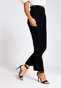 Black high-waisted trousers with pleats, side pockets, and a tailored fit. Paired with strappy black high-heeled sandals.
